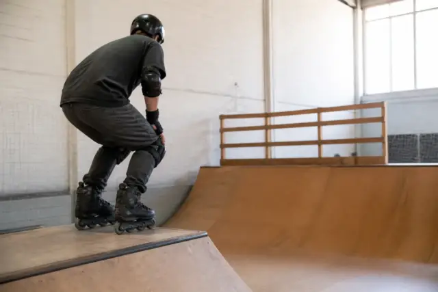 Patineur de profil en protections complètes, genoux fléchis, qui se prépare à descendre une rampe courbe en bois dans un gymnase clair