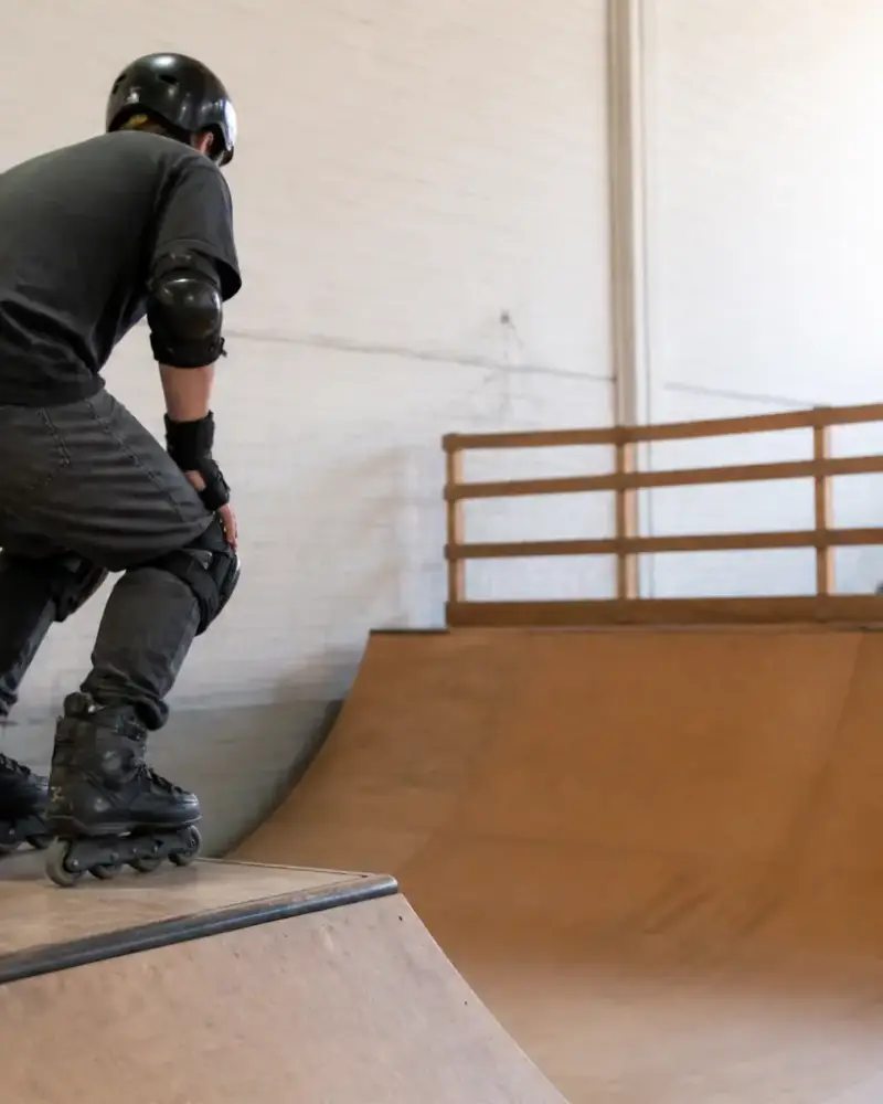 Patineur de profil en protections complètes, genoux fléchis, qui se prépare à descendre une rampe courbe en bois dans un gymnase clair