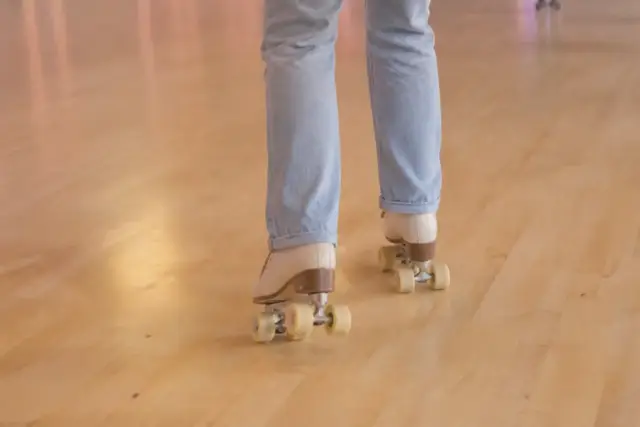 Adulte vu de dos, cadré des genoux aux pieds, chaussé de patins à roulettes quad crème, qui fait ses premiers pas sur un parquet clair