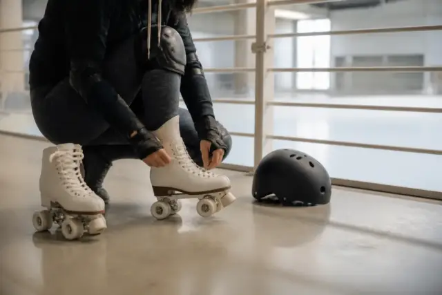 Adulte aux genoux fléchis, casque et protections aux poignets, qui s’appuie sur la rambarde d’un gymnase clair en enfilant ses patins à quatre roues