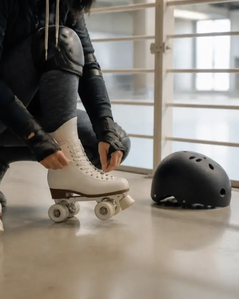 Adulte aux genoux fléchis, casque et protections aux poignets, qui s’appuie sur la rambarde d’un gymnase clair en enfilant ses patins à quatre roues