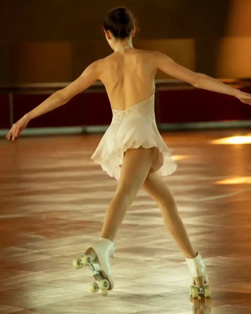 Patineuse en robe claire à jupe courte, bras ouverts, glissant sur un parquet brillant sous une lumière chaude, patins à quatre roues aux roues claires