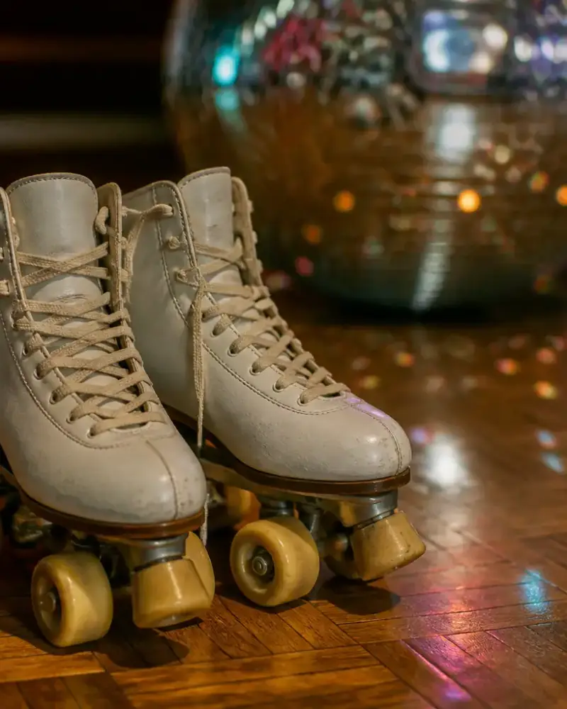 Paire de patins à roulettes à quatre roues aux lacets clairs posée sur un parquet ciré, reflets colorés d’une boule à facettes en arrière-plan