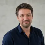 Portrait d’Elouan Lerouet, cheveux châtains courts, barbe légère, chemise bleu nuit, sourire discret, mur gris clair flou en arrière-plan