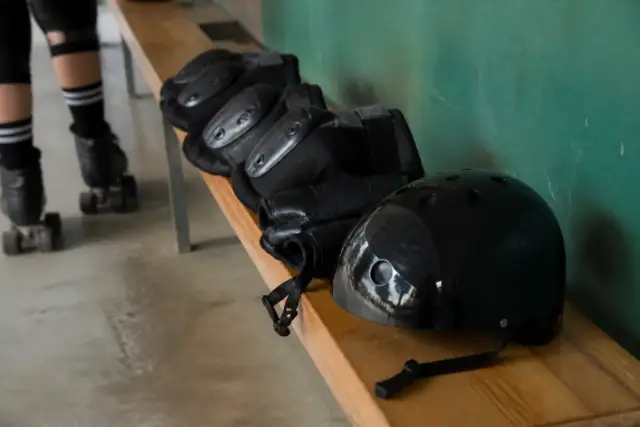 Casque rigide de roller derby sans marque posé sur un banc de bois clair, jugulaire dénouée, genouillères et protège-poignets à côté