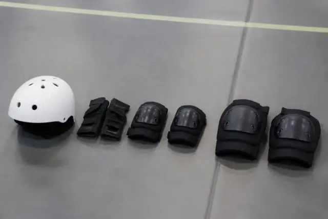 Casque clair, protège-poignets, deux coudières et deux genouillères sans marque disposés en ligne sur un tapis de gymnase gris