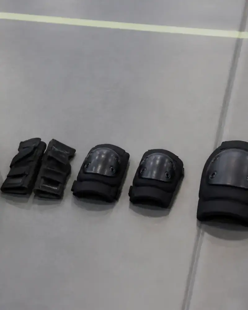 Casque clair, protège-poignets, deux coudières et deux genouillères sans marque disposés en ligne sur un tapis de gymnase gris