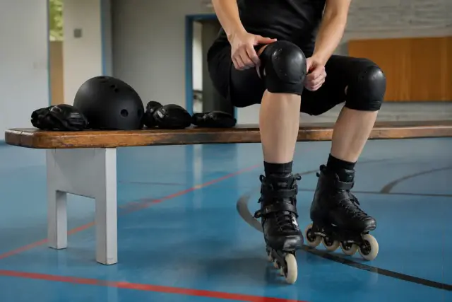 Adulte assis sur un banc de gymnase qui ajuste une genouillère, casque et protège-poignets posés à côté, lumière douce du matin
