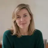 Portrait de Solveig Marvaux, cheveux blonds mi-longs, pull vert sombre, léger sourire, mur crème flou en arrière-plan